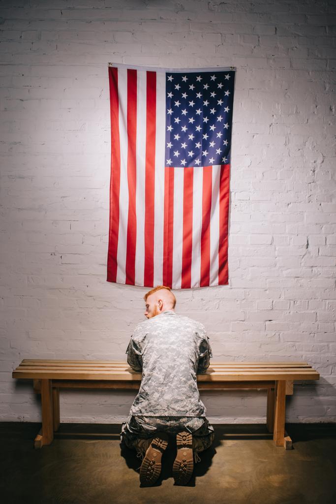 rear view of soldier in military uniform praying with american flag on white brick wall, americas independence day concept - Photo, Image