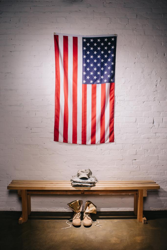 крупным планом виднеется американский флаг, висящий на белой стене и расставленная военная форма на деревянной скамейке
 - Фото, изображение
