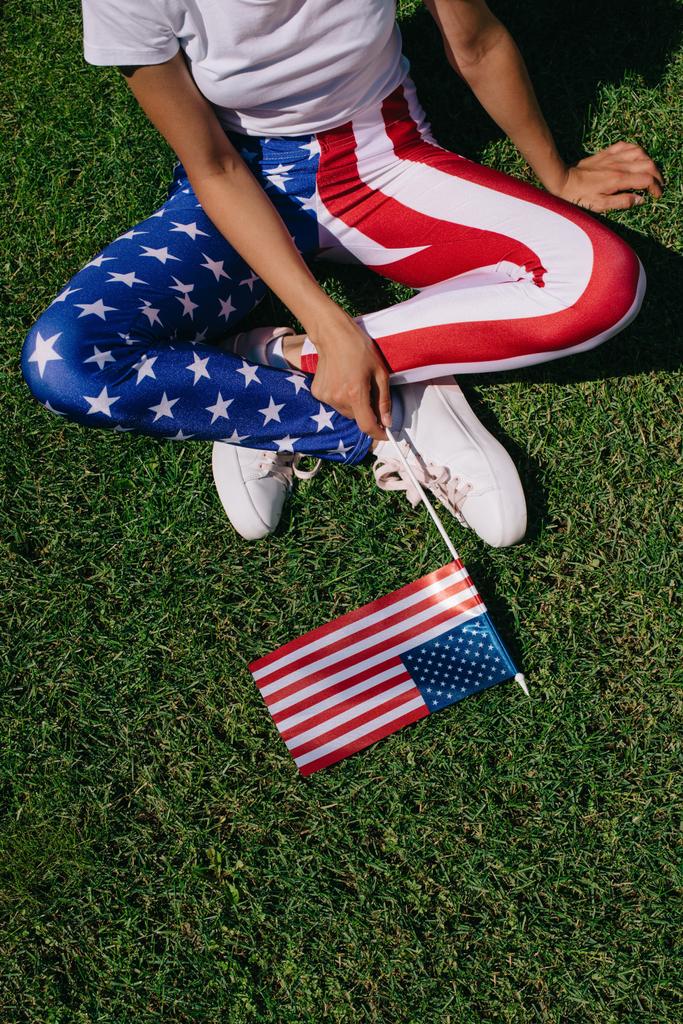 gedeeltelijke weergave van vrouw met vlaggenmast in leggins met Amerikaanse vlag patroon rustend op groene gazon, concept van de vakantie van dag van de onafhankelijkheid van Zuid-Amerika - Foto, afbeelding