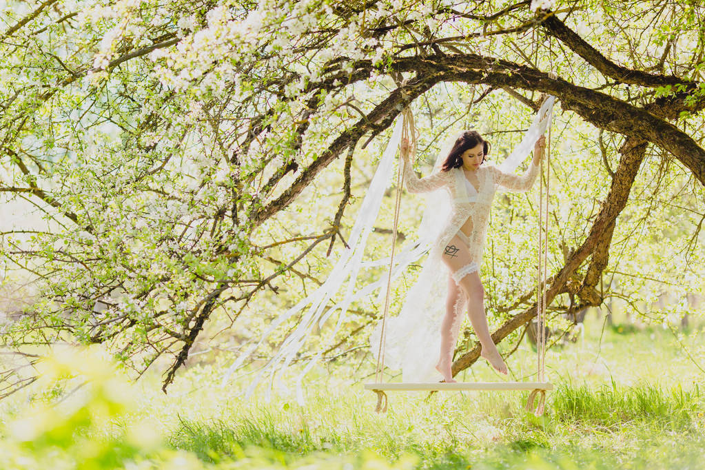 Novia joven seductora en negligee blanco y lencería de pie en un columpio blanco
 - Foto, imagen