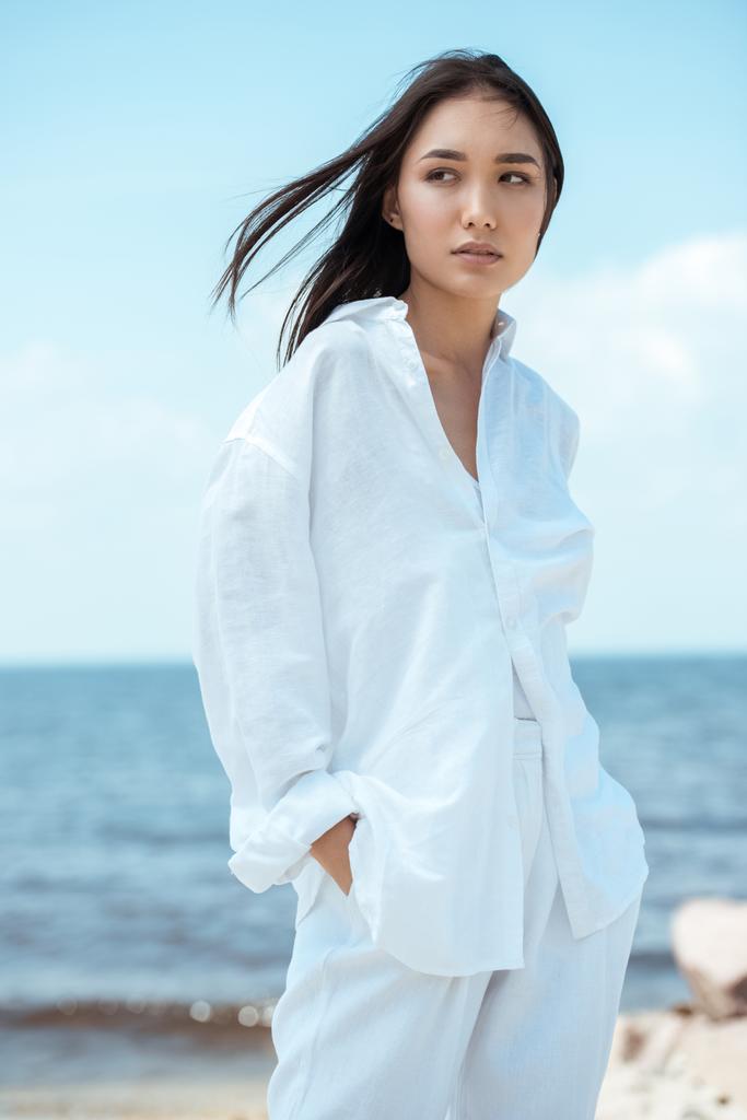 atrakcyjna młoda kobieta azjatyckich z rękami w kieszeniach, patrząc od hotelu na plaży  - Zdjęcie, obraz