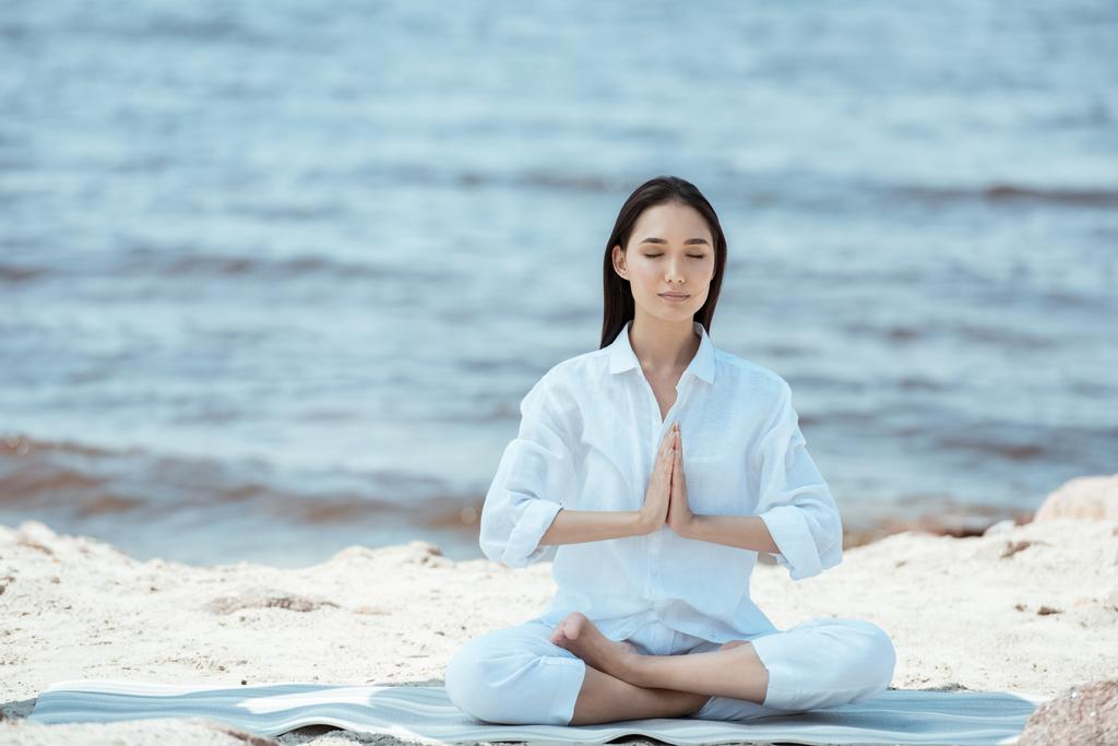 attractive asian woman meditating in anjali mudra (salutation seal) pose on yoga mat by sea  - Photo, Image