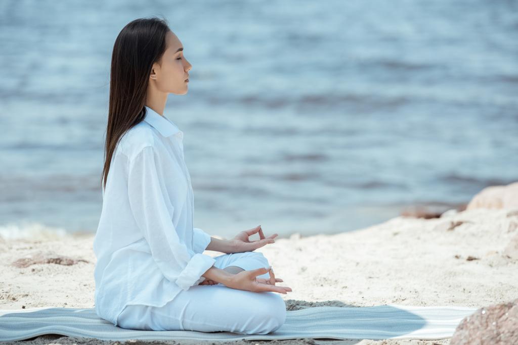 vista laterale della donna focalizzata in ardha padmasana (mezza posa di loto) sul tappetino yoga via mare
  - Foto, immagini