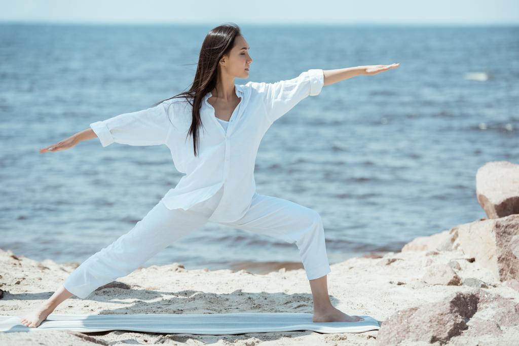 concentrated young asian woman practicing yoga in virabhadrasana (warrior II pose) on mat in front of sea  - Photo, Image
