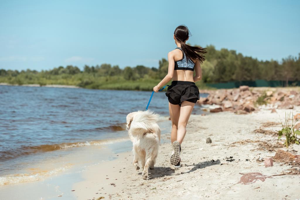 rear view of female jogger running with dog on beach near sea  - Photo, Image