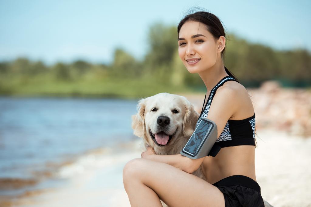 smiling asian sportswoman with smartphone in running armband case embracing golden retriever  - Photo, Image