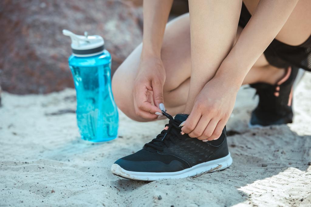 обрезанное изображение спортсменки, завязывающей шнурки возле спортивной бутылки с водой
  - Фото, изображение