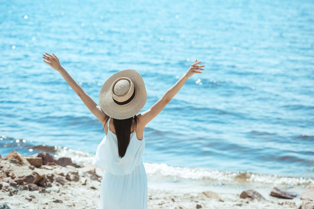 Widok z tyłu z kobieta w białej sukni stojącej z szerokie ramiona przez morze i słomkowy kapelusz  - Zdjęcie, obraz