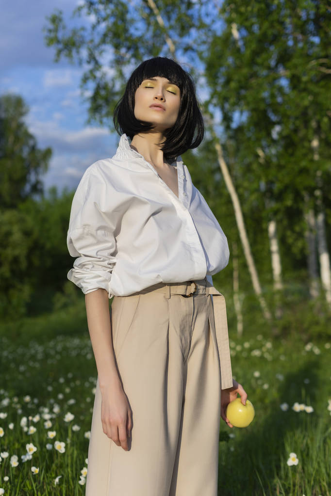 Mooi stijlvol meisje dragen beige broek, een trendy wit overhemd en een zwarte pruik heeft een groene appel in zonlicht op een achtergrond van een blauwe bewolkte hemel. Reclame- en mode-ontwerp. Kopiëren van ruimte. - Foto, afbeelding