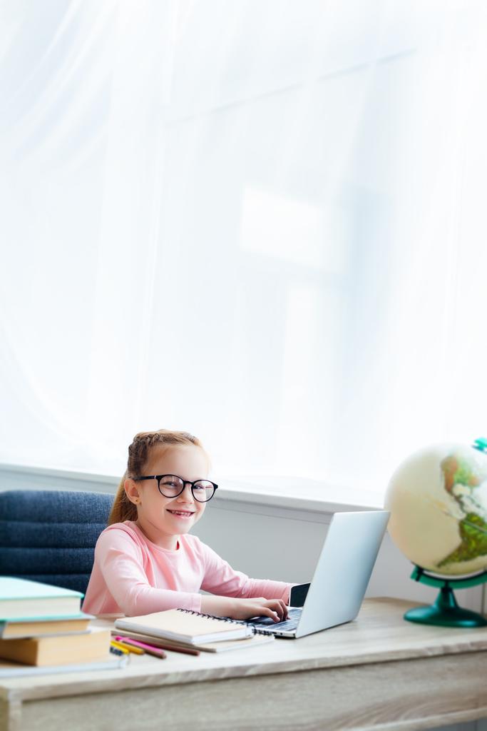 schattig kind in brillen glimlachend in de camera terwijl het bestuderen met laptop thuis - Foto, afbeelding