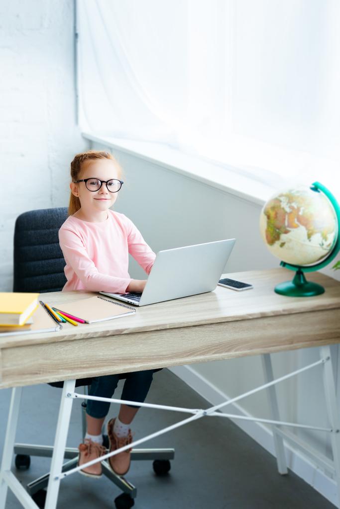 Hochwinkelbild eines Kindes in Brille, das mit Laptop in die Kamera lächelt und zu Hause lernt - Foto, Bild