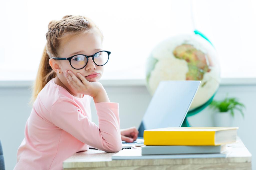 gelangweiltes Kind mit Brille blickt beim Lernen mit Laptop und Büchern in die Kamera - Foto, Bild