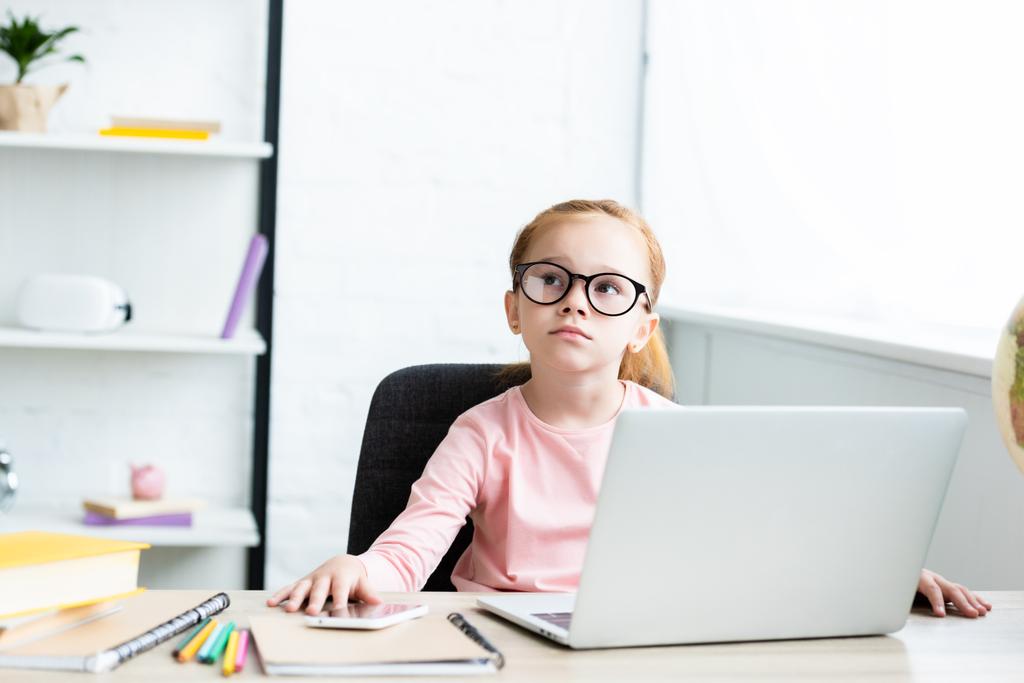 Nachdenkliches kleines Schulkind mit Brille blickt auf, während es am Schreibtisch sitzt und Laptop benutzt - Foto, Bild