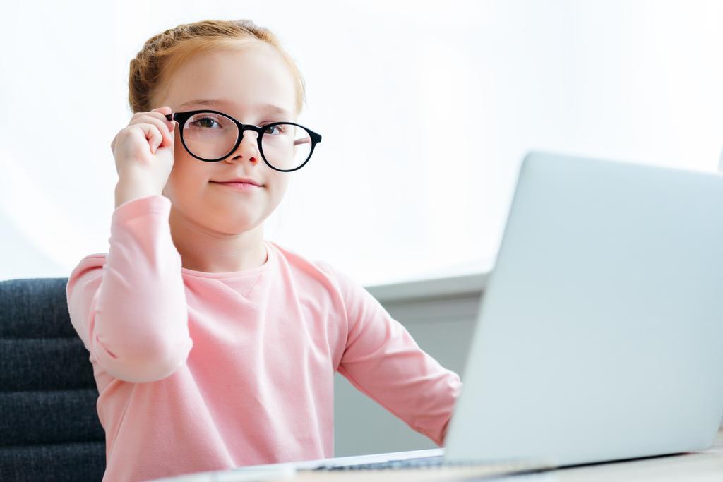 beautiful schoolchild adjusting eyeglasses and smiling at camera while using laptop - Photo, Image