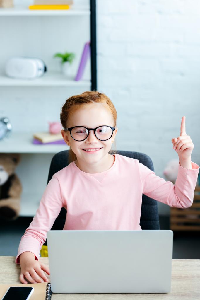αξιολάτρευτο κόκκινα μαλλιά μαθήτρια σε γυαλιά στραμμένο προς τα επάνω με το δάχτυλο και χαμογελαστός στην κάμερα ενώ χρησιμοποιείτε φορητό υπολογιστή στο σπίτι - Φωτογραφία, εικόνα