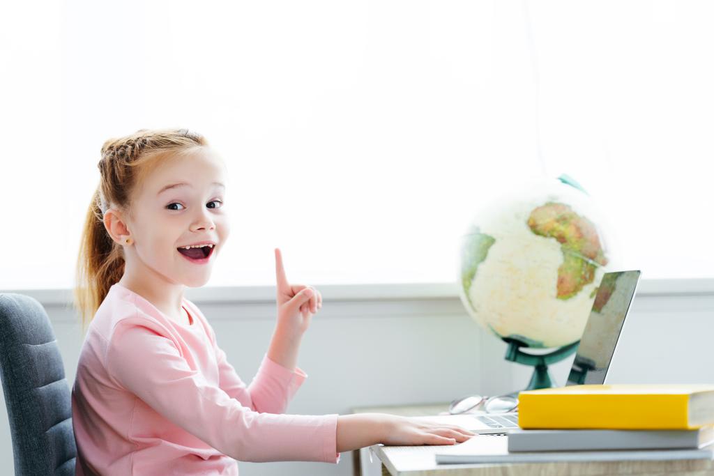 alegre pelirroja niño apuntando hacia arriba con el dedo y sonriendo a la cámara mientras estudia con libros y portátil en casa
 - Foto, imagen