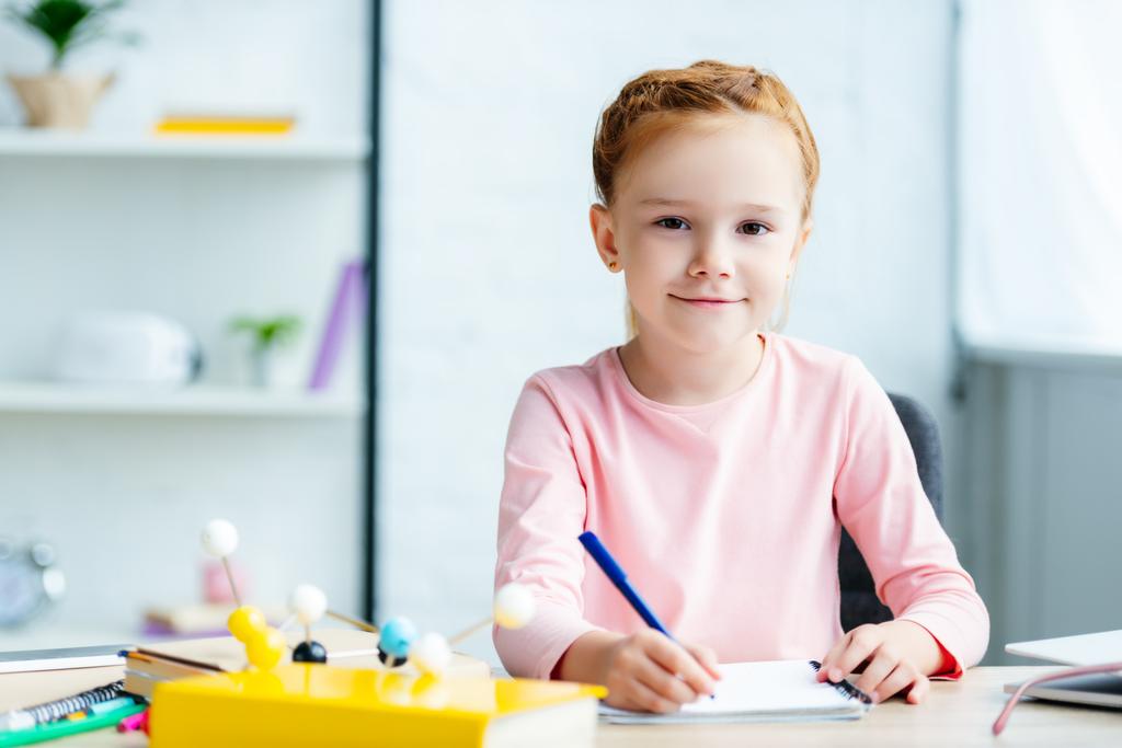 gyönyörű vörös hajú iskolás mosolyogva kamera írásakor a notebook-on iskolapad - Fotó, kép