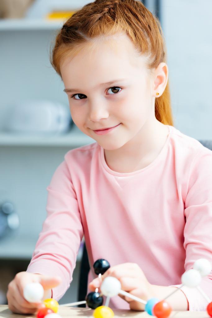 mooie roodharige schoolmeisje holding moleculair model en glimlachend op camera - Foto, afbeelding