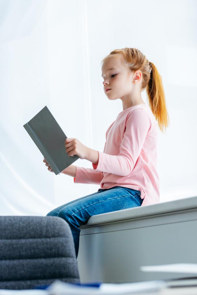 χαμηλή γωνία θέα της χαριτωμένο μικρό μαθήτρια κάθεται στο περβάζι και ανάγνωση βιβλίων - Φωτογραφία, εικόνα