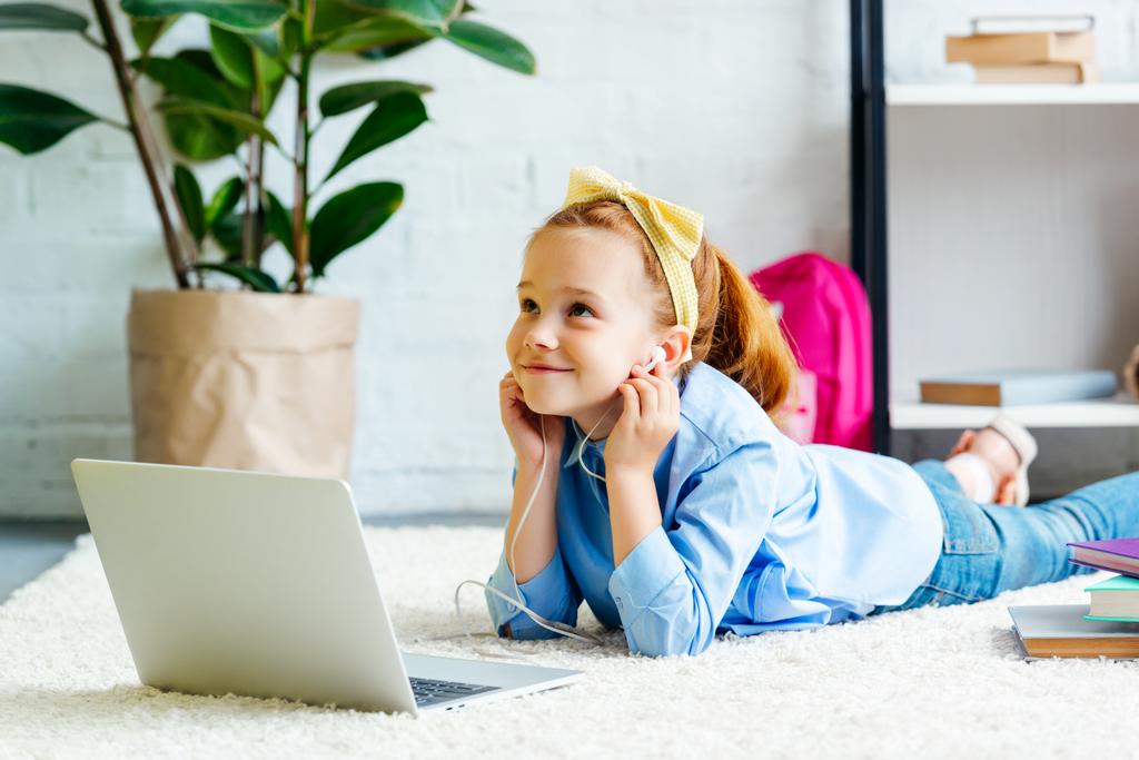 Nachdenklich lächelndes Kind mit Kopfhörern, das aufschaut, während es auf dem Teppich liegt und zu Hause Laptop benutzt - Foto, Bild
