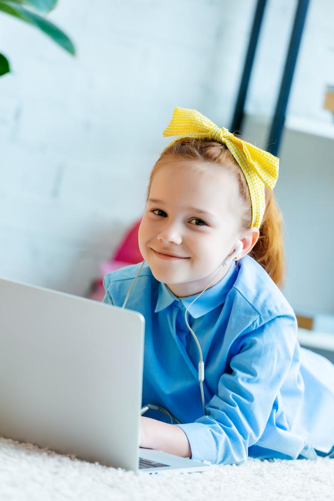 schöne rothaarige Kind mit Kopfhörern lächelt in die Kamera, während auf dem Teppich liegend und mit Laptop  - Foto, Bild