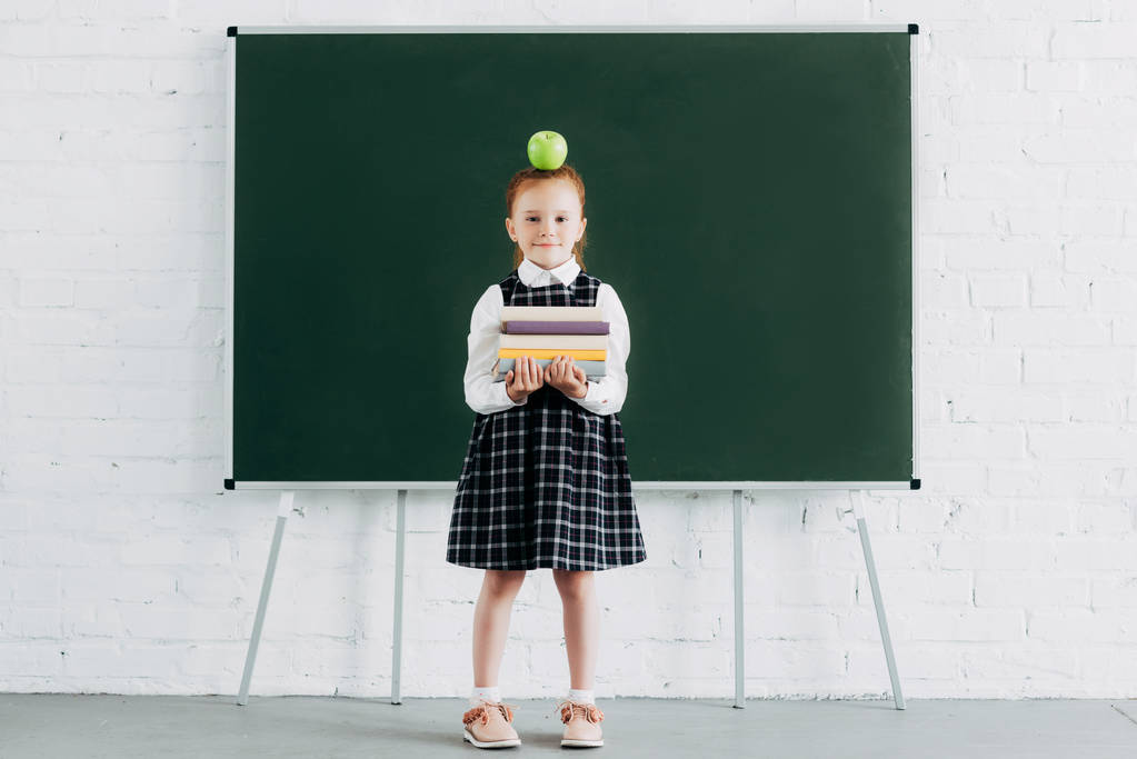 cute little schoolgirl with apple on head holding pile of books and looking at camera while standing near blackboard - Photo, Image