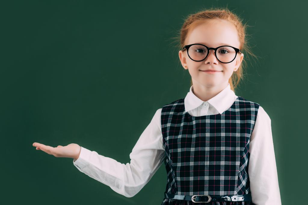 adorable little schoolgirl in eyeglasses showing blank chalkboard and smiling at camera  - Photo, Image
