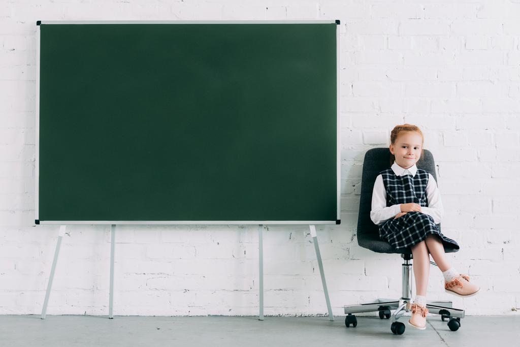 schattige kleine schoolmeisje glimlachend op camera zittend op een stoel in de buurt van leeg schoolbord - Foto, afbeelding