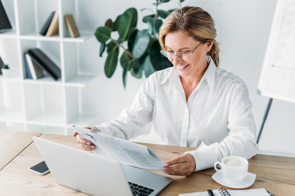 sorridente attraente donna d'affari che legge il giornale a tavola con computer portatile in ufficio
 - Foto, immagini