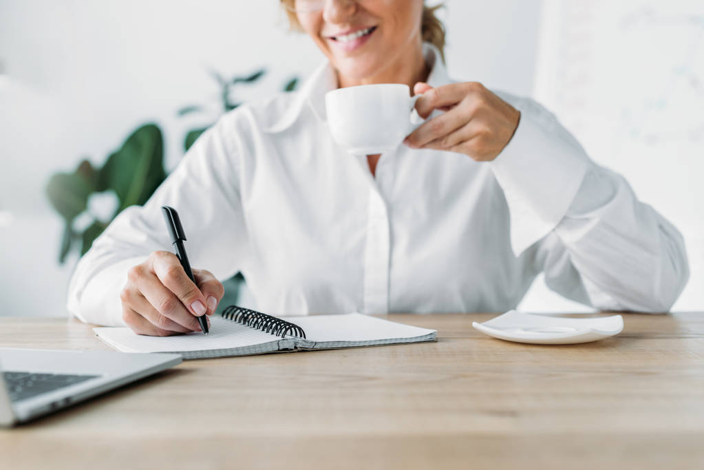 bijgesneden afbeelding zakenvrouw kopje koffie houden en het schrijven naar notebook in kantoor - Foto, afbeelding