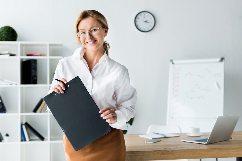 smiling attractive businesswoman holding clipboard and looking at camera in office - Photo, Image
