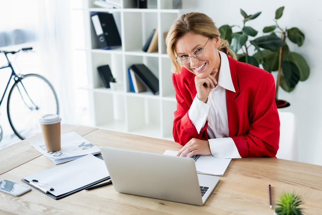 привлекательная улыбающаяся деловая женщина сидит за столом с ноутбуком в офисе и смотрит в камеру
 - Фото, изображение