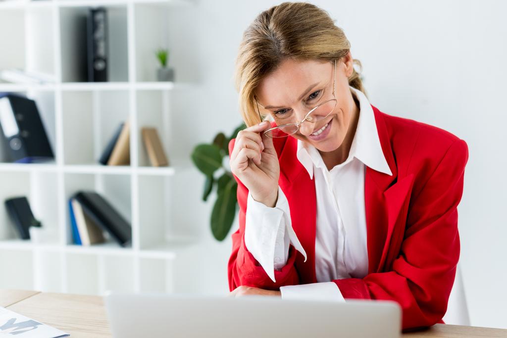 sorridente atraente empresária tocando óculos e olhando para laptop no escritório
 - Foto, Imagem