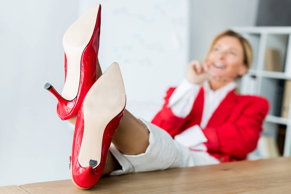 привлекательная деловая женщина смеется и сидит с ногами на столе в офисе в красной обуви на переднем плане
 - Фото, изображение