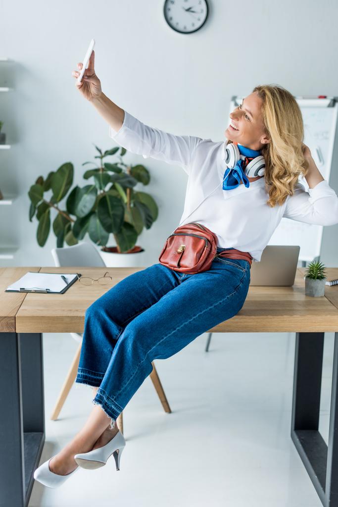 atraktivní podnikatelka sedí na stole v kanceláři a pořizování selfie s smartphone - Fotografie, Obrázek