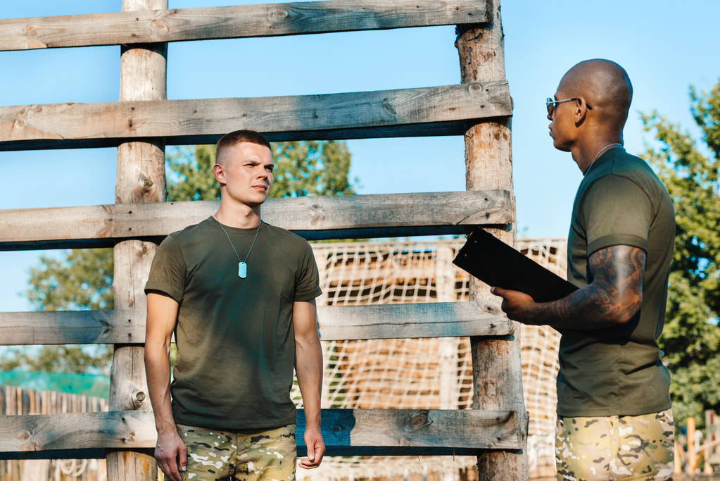 instructor táctico afroamericano con bloc de notas y joven soldado en uniforme militar en rango
 - Foto, Imagen