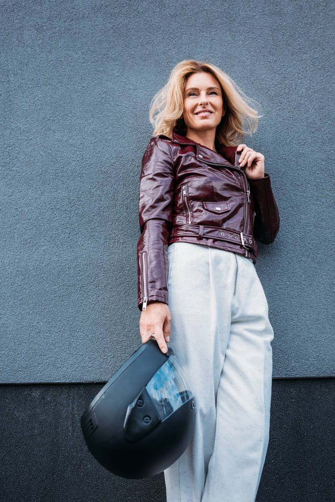 lage hoekmening van lachende vrouw in leren jas Motorhelm houden op straat en op zoek weg - Foto, afbeelding