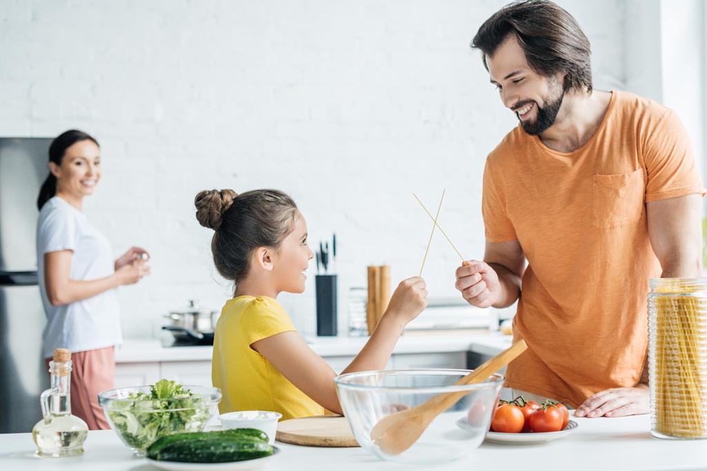 vader en dochter spelen met stokjes terwijl moeder permanent wazig op achtergrond op keuken - Foto, afbeelding