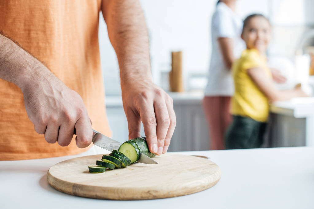 rajattu laukaus isä leikkaa kurkkua salaattia, kun hänen vaimonsa ja tyttärensä tiskaavat keittiössä
 - Valokuva, kuva