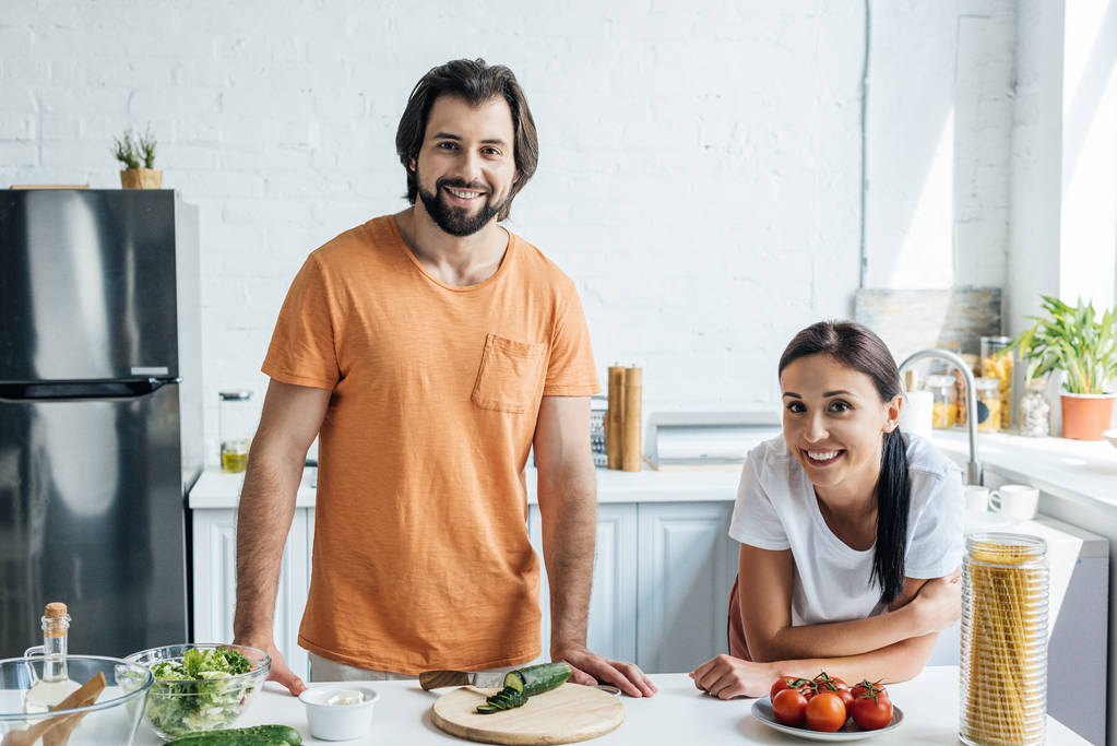 hermosa pareja de adultos mirando a la cámara mientras preparan ensalada juntos en la cocina
 - Foto, imagen