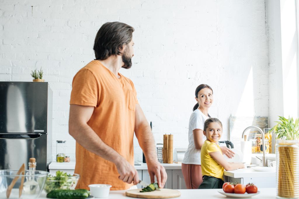 attraente giovane padre taglio cetriolo per insalata mentre la moglie e la figlia lavare i piatti in cucina
 - Foto, immagini