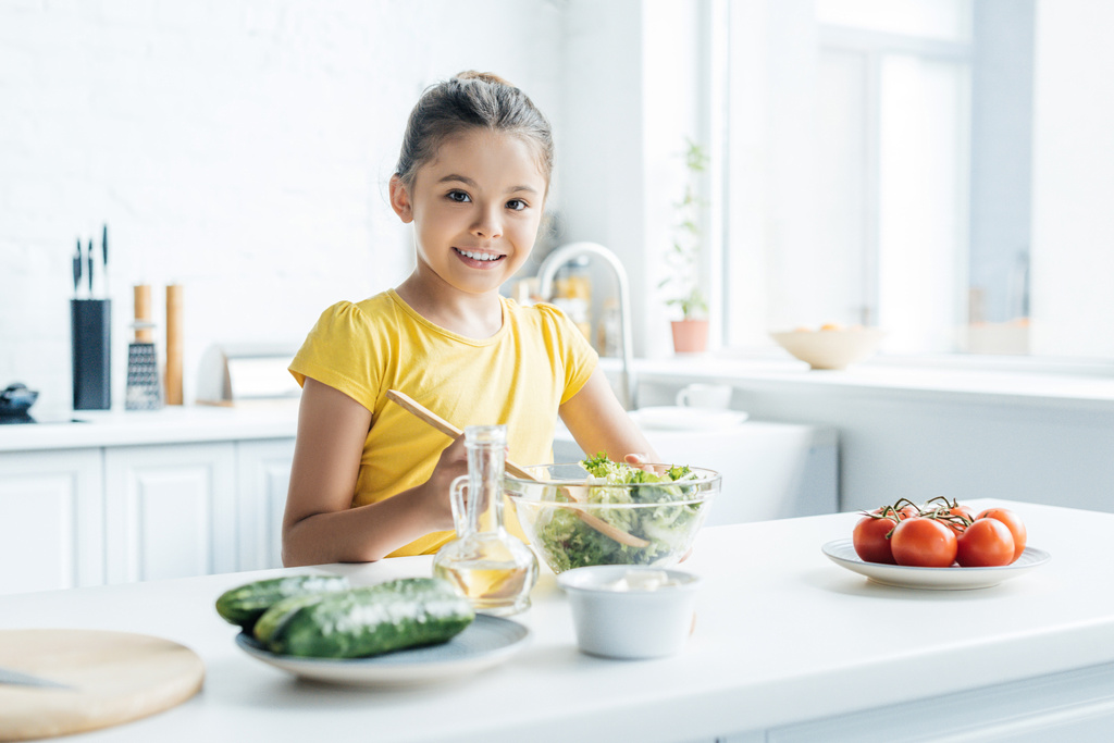 mosolygós kislány készül a saláta, a konyha és a látszó-on fényképezőgép - Fotó, kép