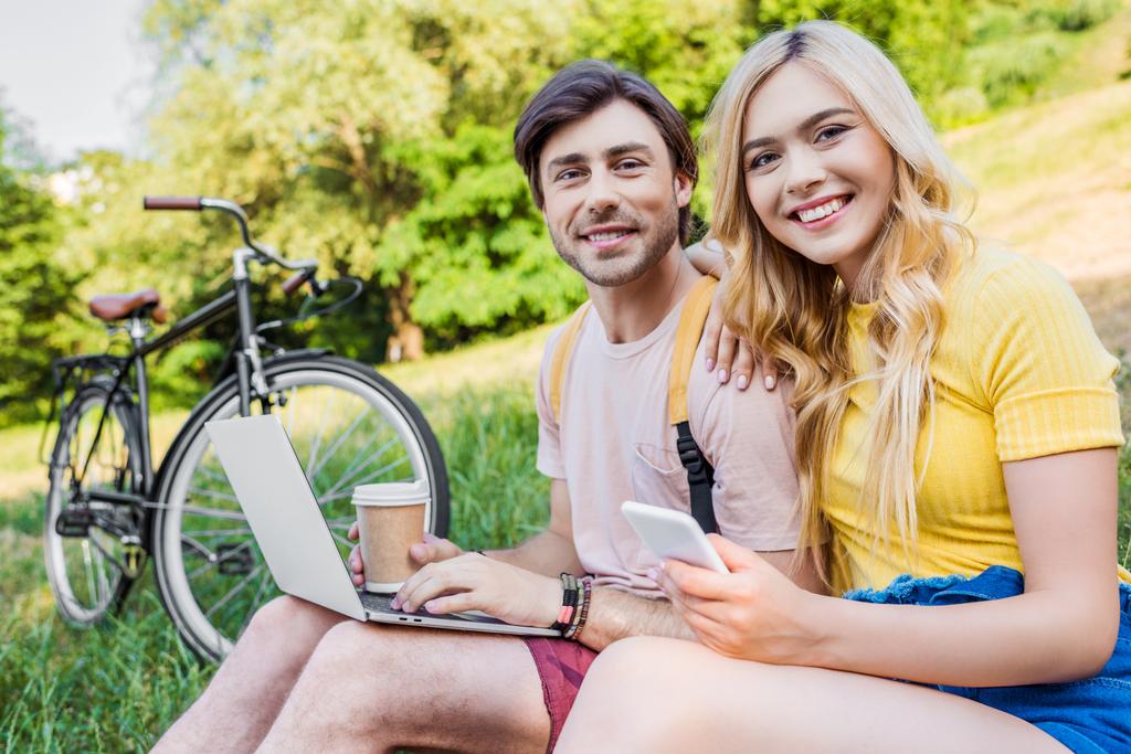 sonriente pareja con portátil y teléfono inteligente descansando sobre hierba verde en el parque
 - Foto, imagen