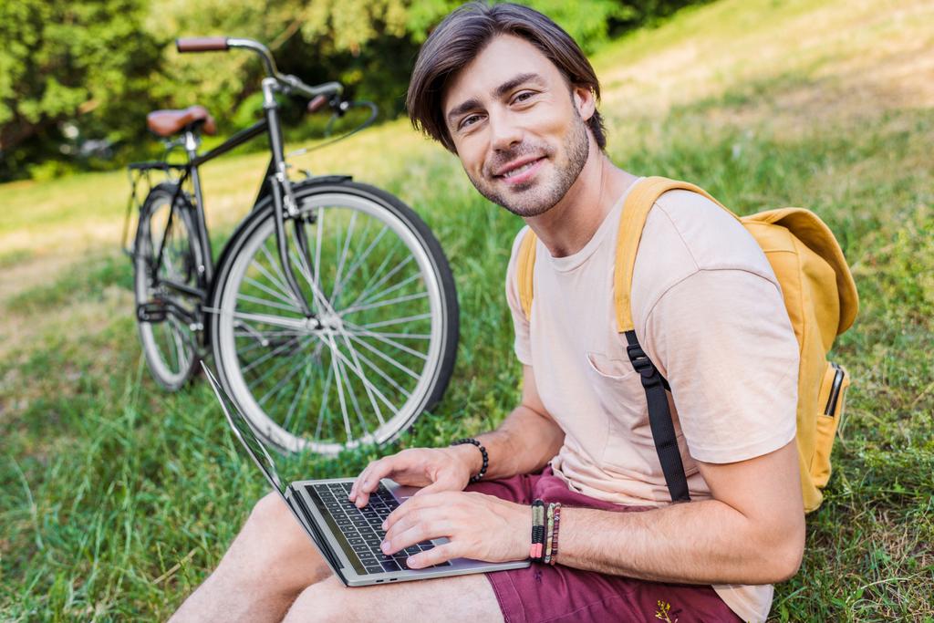 widok boczny uśmiechnięty człowiek z plecaka, laptop i rowerów w pobliżu siedząc na zielonej trawie w parku - Zdjęcie, obraz