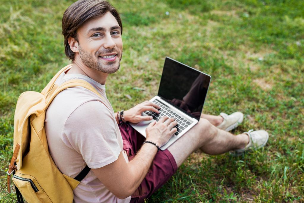 Zijaanzicht van glimlachende man met rugzak en laptop zitting op groen gras in park - Foto, afbeelding