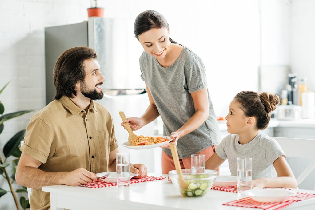 attraktive junge Mutter mit leckerem Frühstück für Tochter und Ehemann in der Küche - Foto, Bild