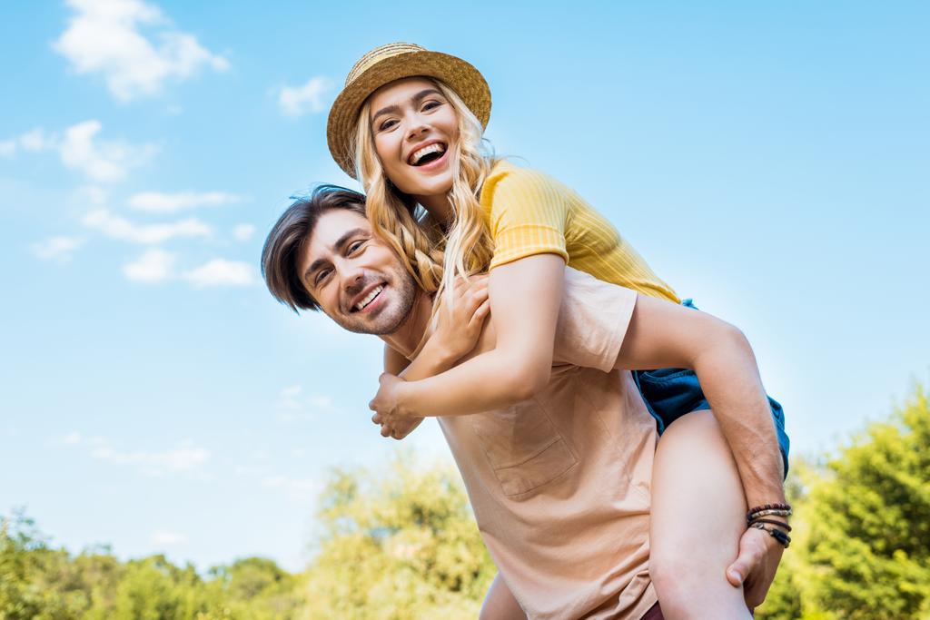 χαμηλή γωνία προβολής του ευτυχισμένο ζευγάρι piggybacking στο πάρκο - Φωτογραφία, εικόνα