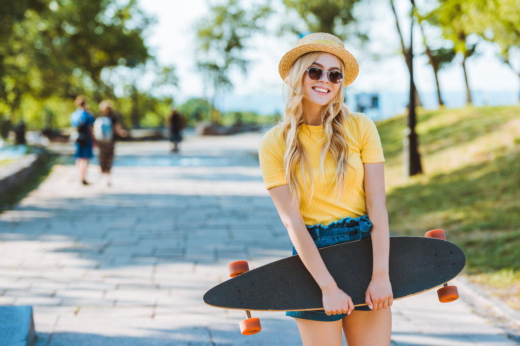 portret uśmiechający się blond kobieta w okulary przeciwsłoneczne i kapelusz z longboard w ręce na ulicy - Zdjęcie, obraz