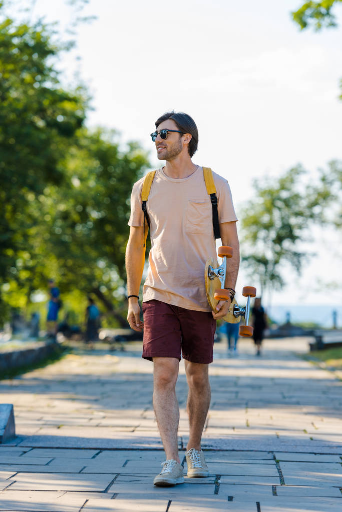 молодой человек в солнцезащитных очках с рюкзаком и лонгбордом ходит по улице
 - Фото, изображение