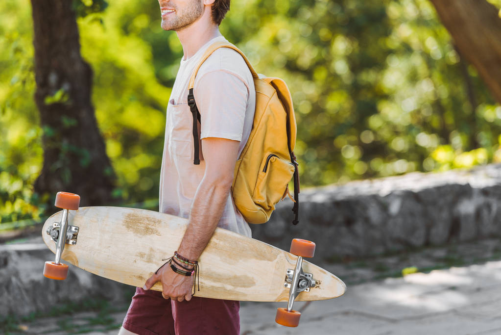 vista parcial del hombre sonriente con mochila y longboard en la calle
 - Foto, imagen